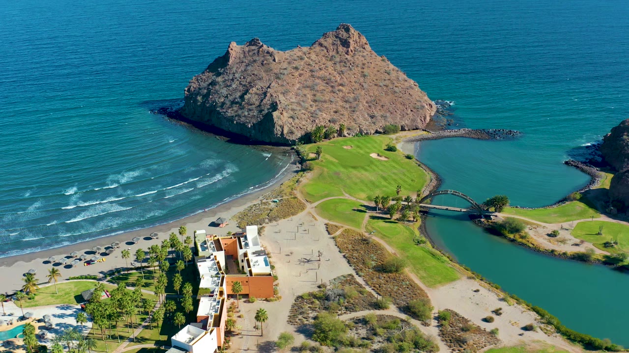 A high drone view in Loreto, Baja California Sur keeps the rocky outcrop centered above the beach while small wave lines move toward shore and no visible people appear.