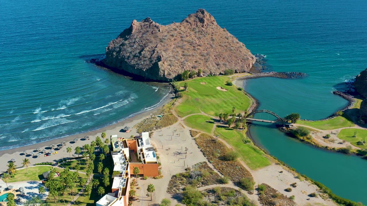 The Loreto aerial composition shows turquoise water on both sides of the beach, the rocky island at the top, a bridge crossing the lagoon, nearby buildings, and no visible people.