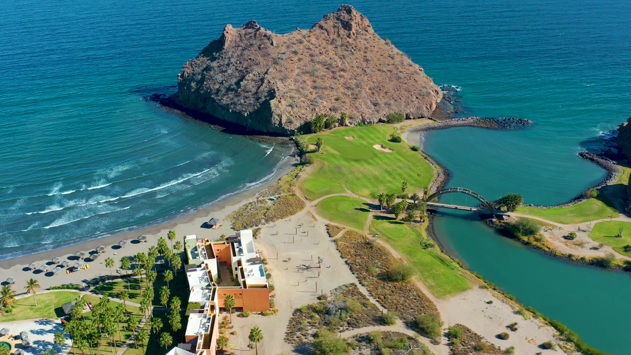 Rocky island above beach and lagoon with bridge and coastal buildings below.