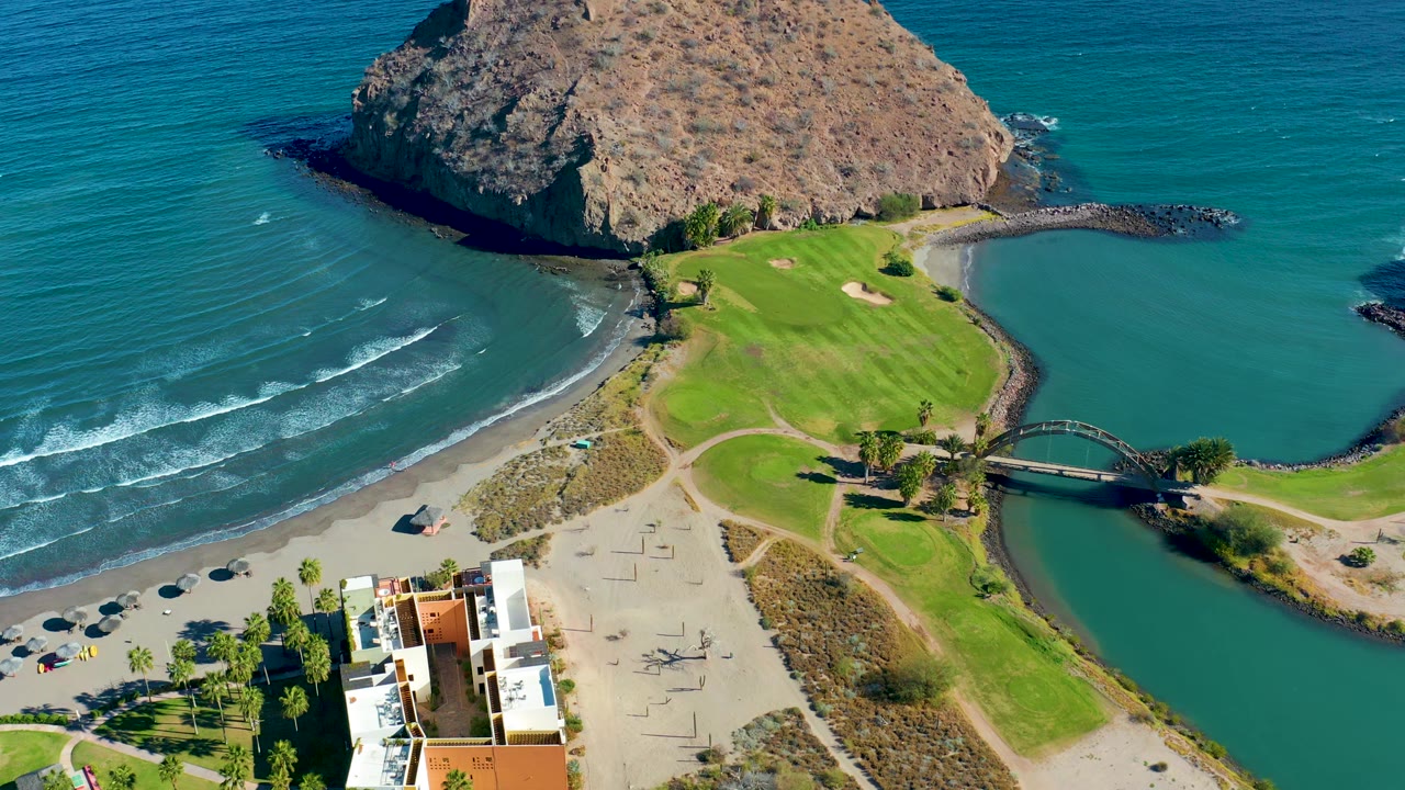 Second aerial frame over Loreto shows small wave lines rolling toward the beach, fairways below the rocky headland, the bridge crossing the inlet, and no visible people.
