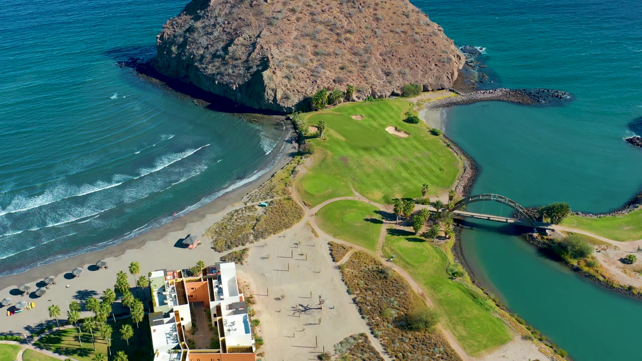At 0:02 the Loreto aerial view holds the rocky headland across the top, a green course beside the inlet, beach umbrellas at lower left, and no visible people.
