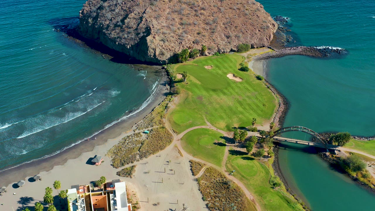Aerial frame over Loreto, Baja California Sur showing teal ocean, white surf lines, sandy beach, green golf course, curved bridge, and rocky headland with no visible people.