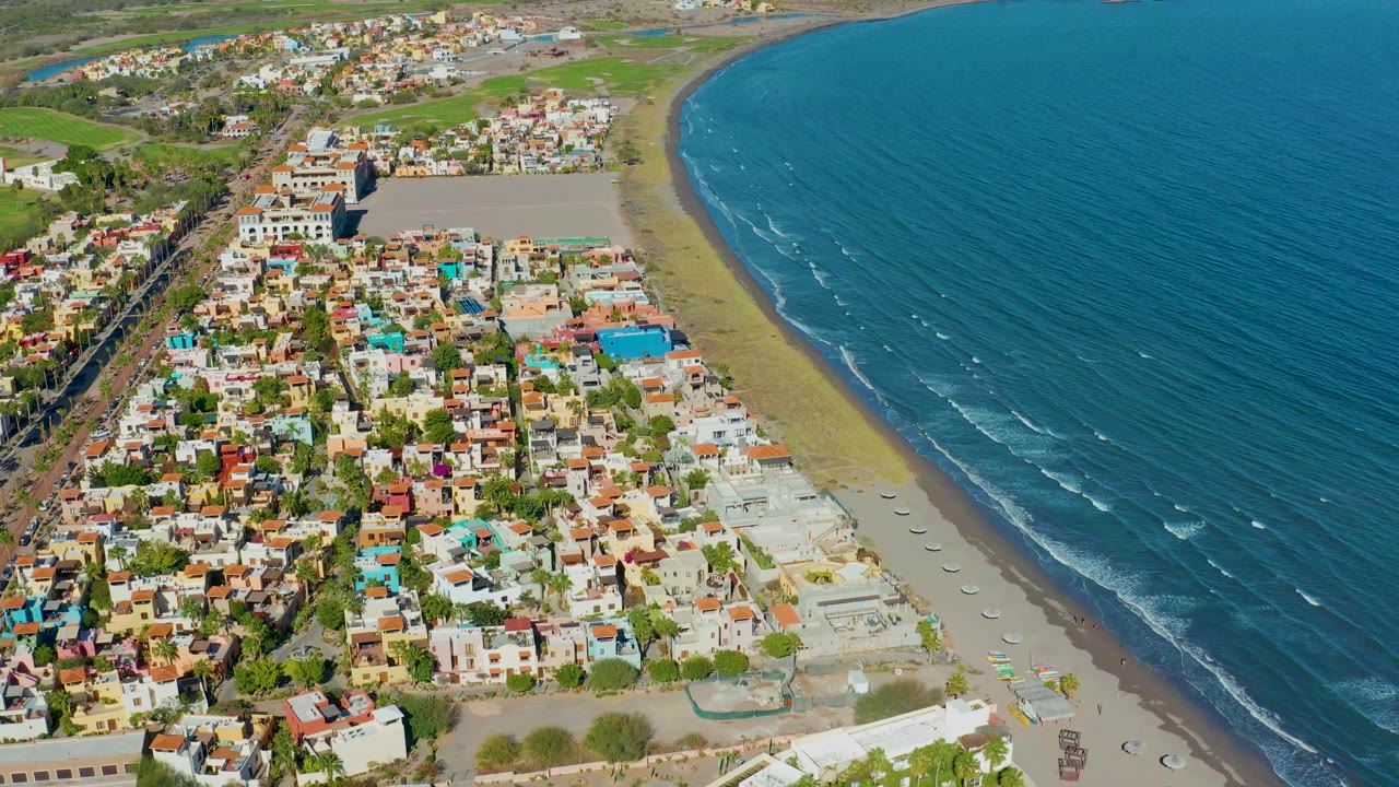 Aerial view over Loreto with dense residential blocks at left, a pale sandy beach in the center, ocean waves slightly farther along the shore, and no visible people.