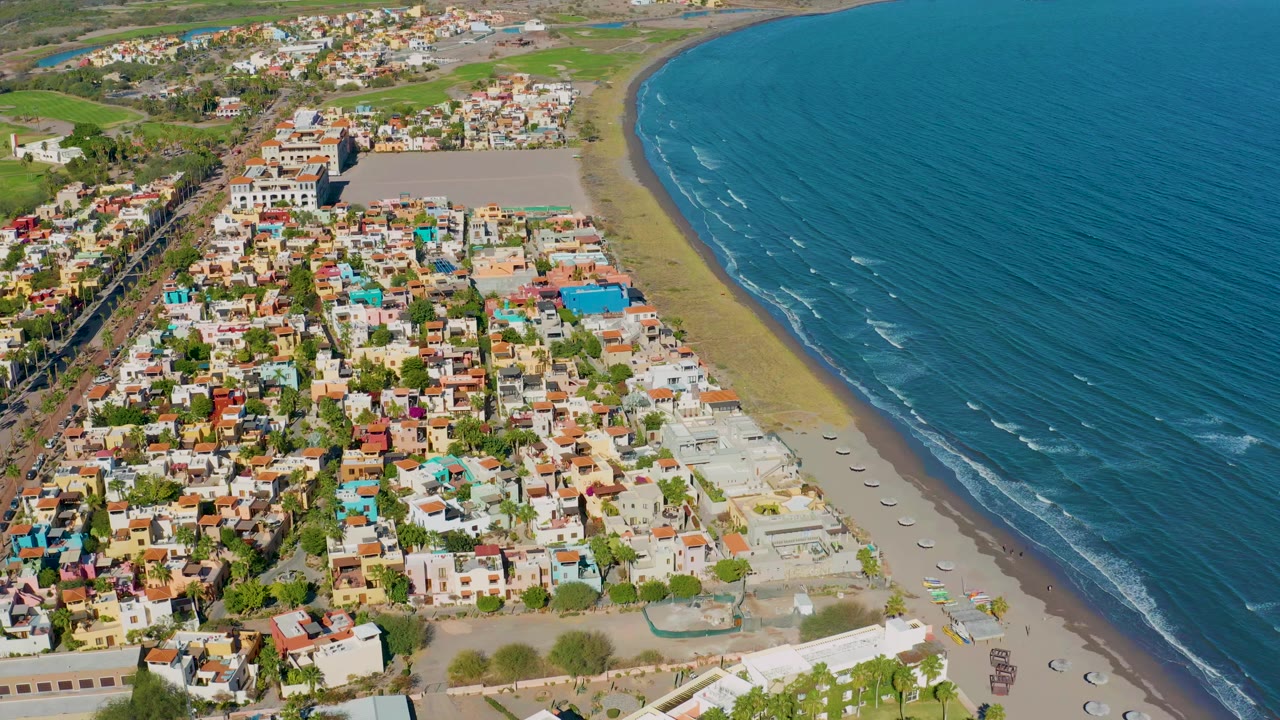 High aerial view of Loreto, Baja California Sur, with colorful low-rise homes beside a curving sandy beach and blue ocean water, no visible people.