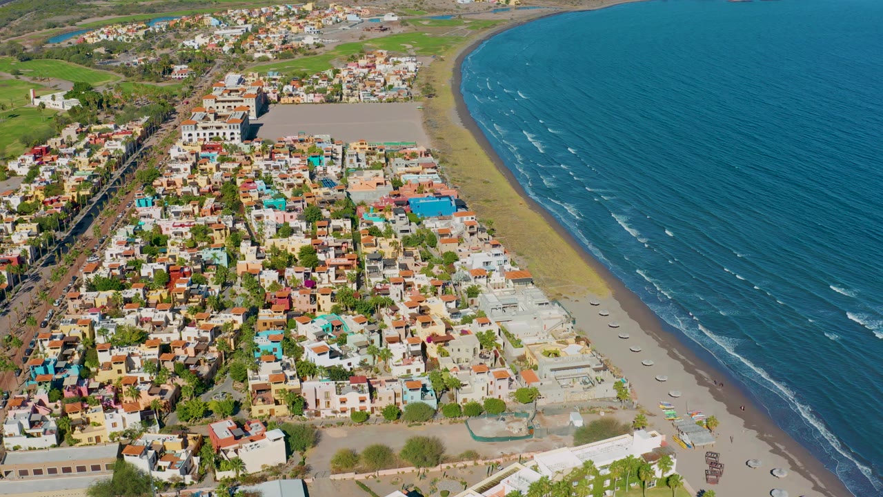 Aerial frame over Loreto with houses and streets filling the left side, the beach narrowing toward the upper frame, ocean waves on the right, and no visible people.