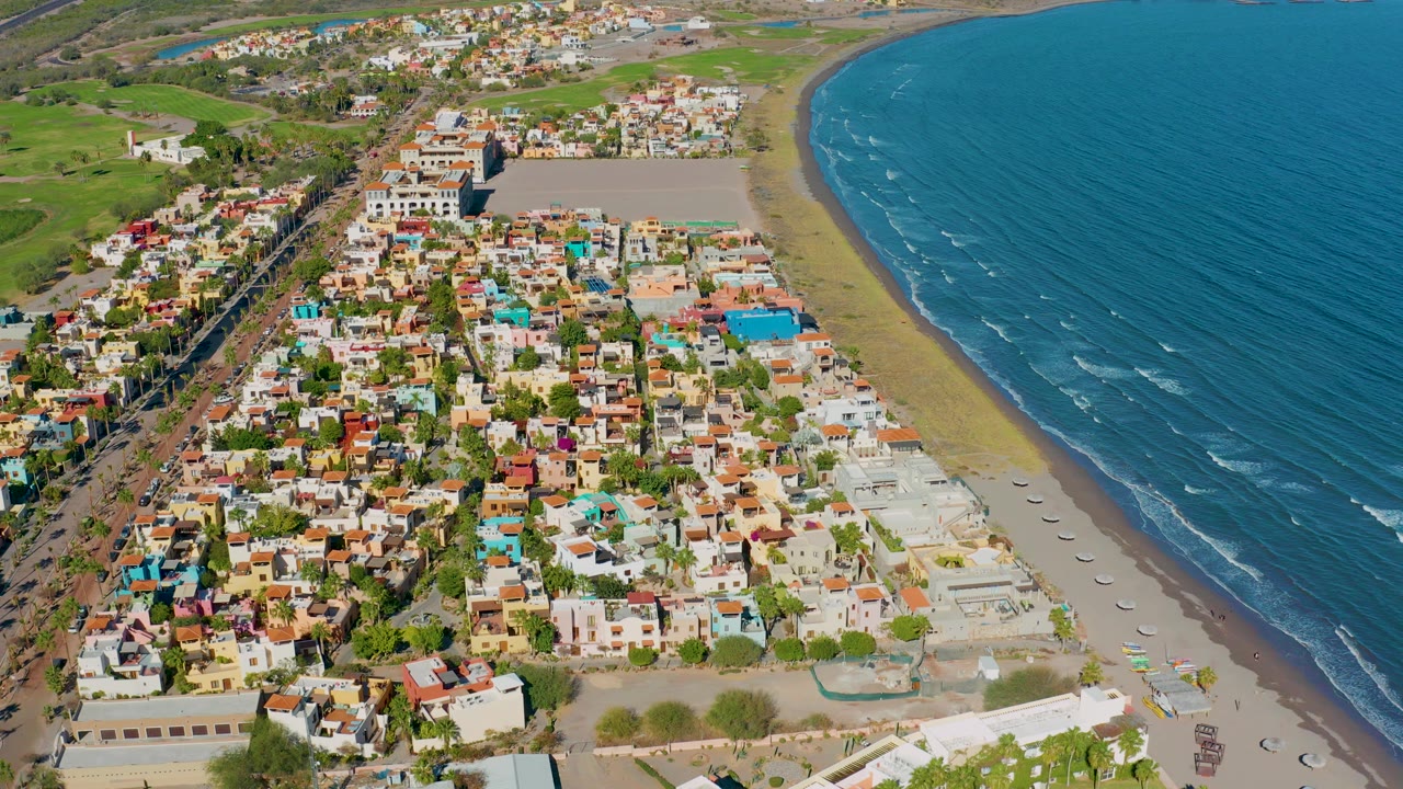 High aerial view over Loreto, Baja California Sur, with colorful low buildings beside a curved sandy beach, blue ocean to the right, green inland fields beyond, and no visible people.