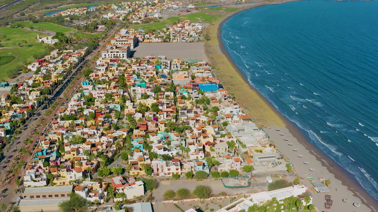 High drone perspective over Loreto with the town filling the middle of frame, the beach curving along the right, blue ocean occupying the upper right, inland greenery at left, and no visible people.