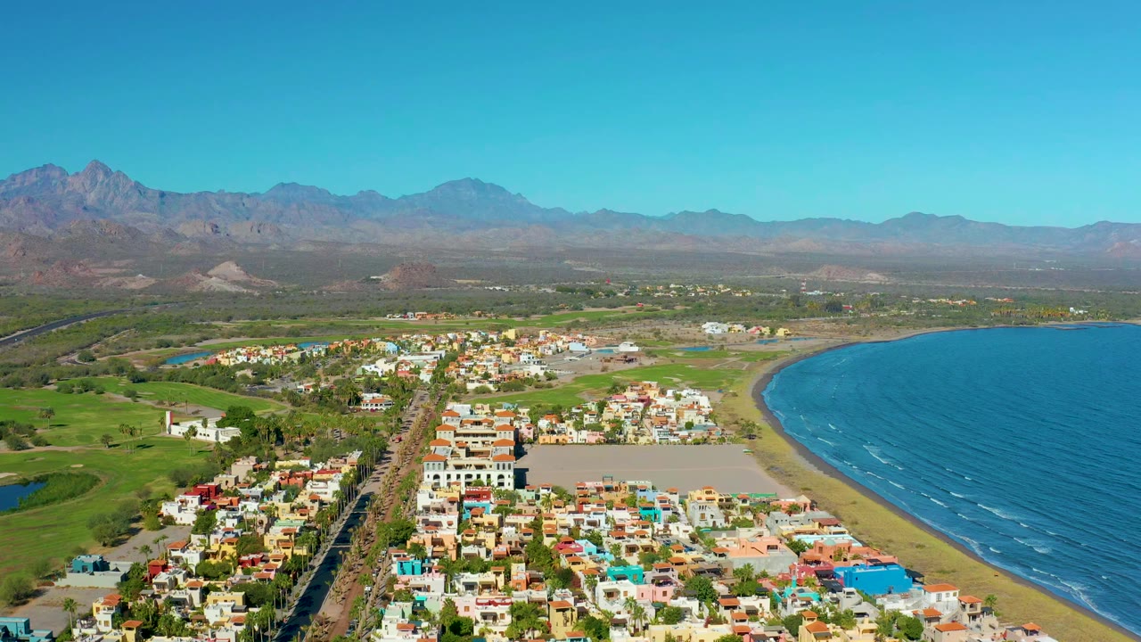 Aerial view over Loreto, Baja California Sur, showing colorful town blocks, a straight road, green fields, a curved beach, blue water, distant mountains, and no visible people.