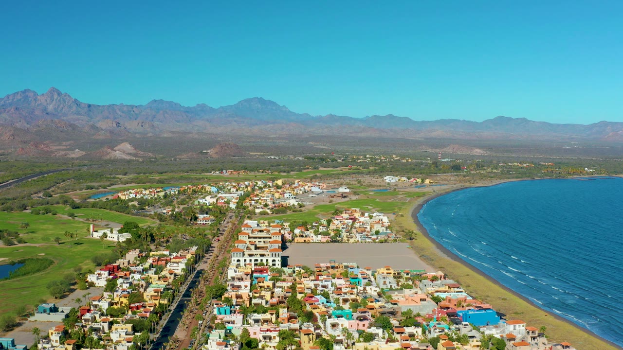 Aerial shoreline view with road, green fields, town buildings, ocean, and mountains.