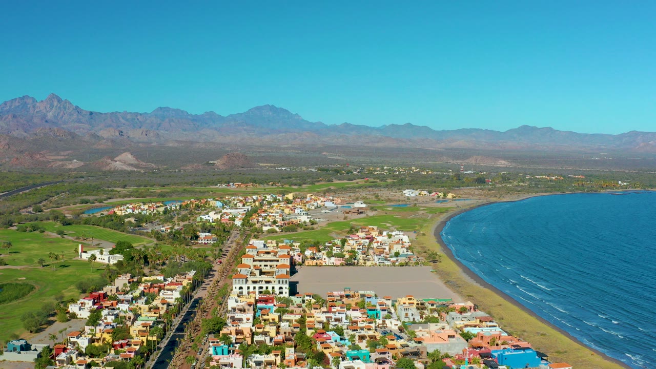 Opening aerial frame over Loreto, Baja California Sur, with colorful buildings in the foreground, a sandy beach and blue ocean to the right, distant mountains under clear sky, and no visible people.