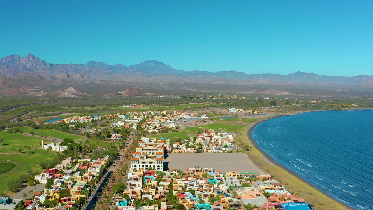 A wide 4K aerial frame of Loreto with the town and straight road below, the sandy beach curving beside blue ocean on the right, green land inland, distant mountains, and no visible people.