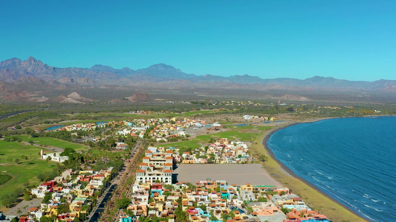 Aerial view over beachside buildings, inland fields, ocean, and mountains.