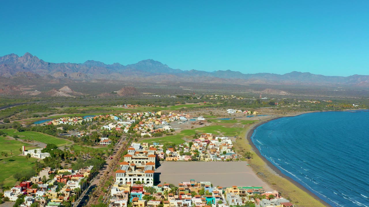 Wide aerial frame over Loreto, Baja California Sur, showing low buildings, a curved sandy beach, blue coastal water, green fields, distant mountains, and no visible people.