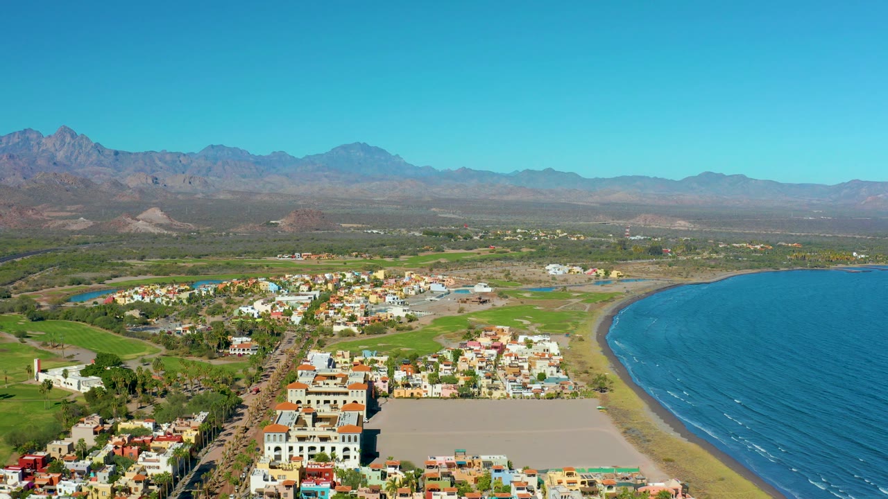 Aerial coastal town scene with road, beach, fields, and mountain backdrop.