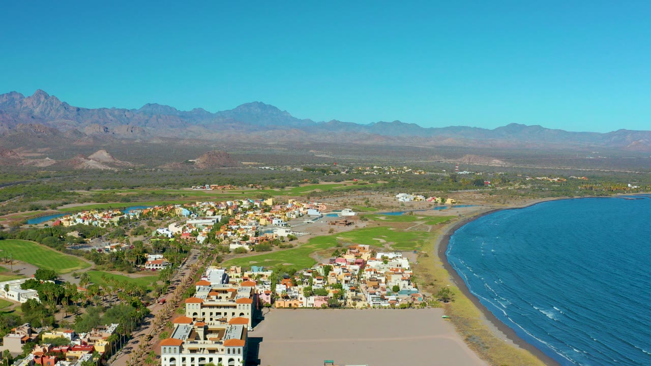 Aerial opening view over Loreto, Baja California Sur, with a sandy beach and blue ocean at right, low buildings near shore, green fields inland, distant mountains, and no visible people.