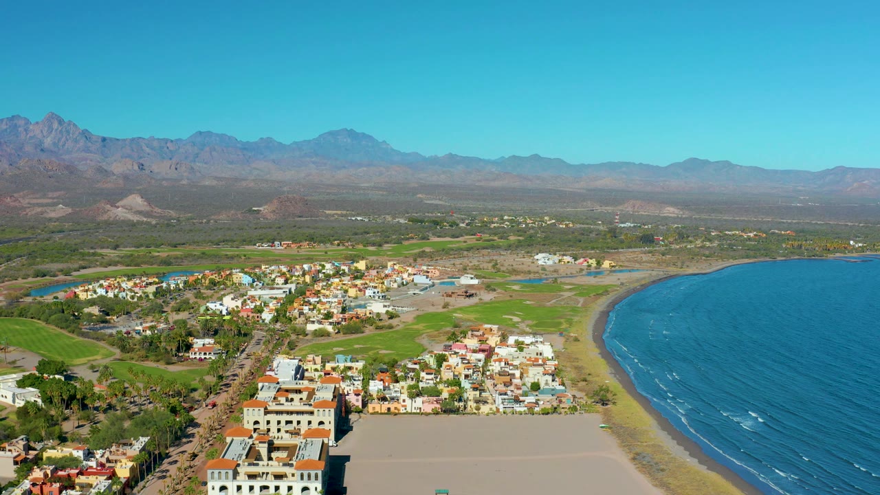Aerial view of Loreto with the curved sandy shore and blue water more prominent on the right, clustered homes and streets in the center, green inland fields, mountains, and no visible people.