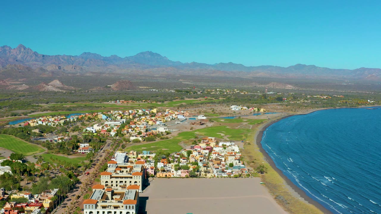 Aerial view of beachside buildings, green fields, ocean, and mountains under clear sky.