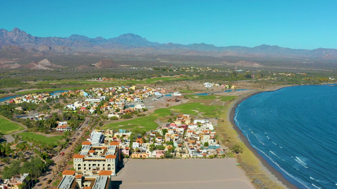 Aerial view over Loreto, Baja California Sur, showing low-rise town blocks beside a sandy beach, blue ocean to the right, green fields inland, distant mountains, and no visible people.