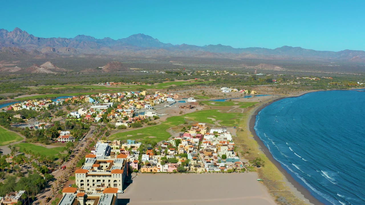 Aerial view across Loreto's beachside neighborhood, with dense light-colored buildings near the sand, blue water on the right, green fields behind town, rugged mountains, and no visible people.