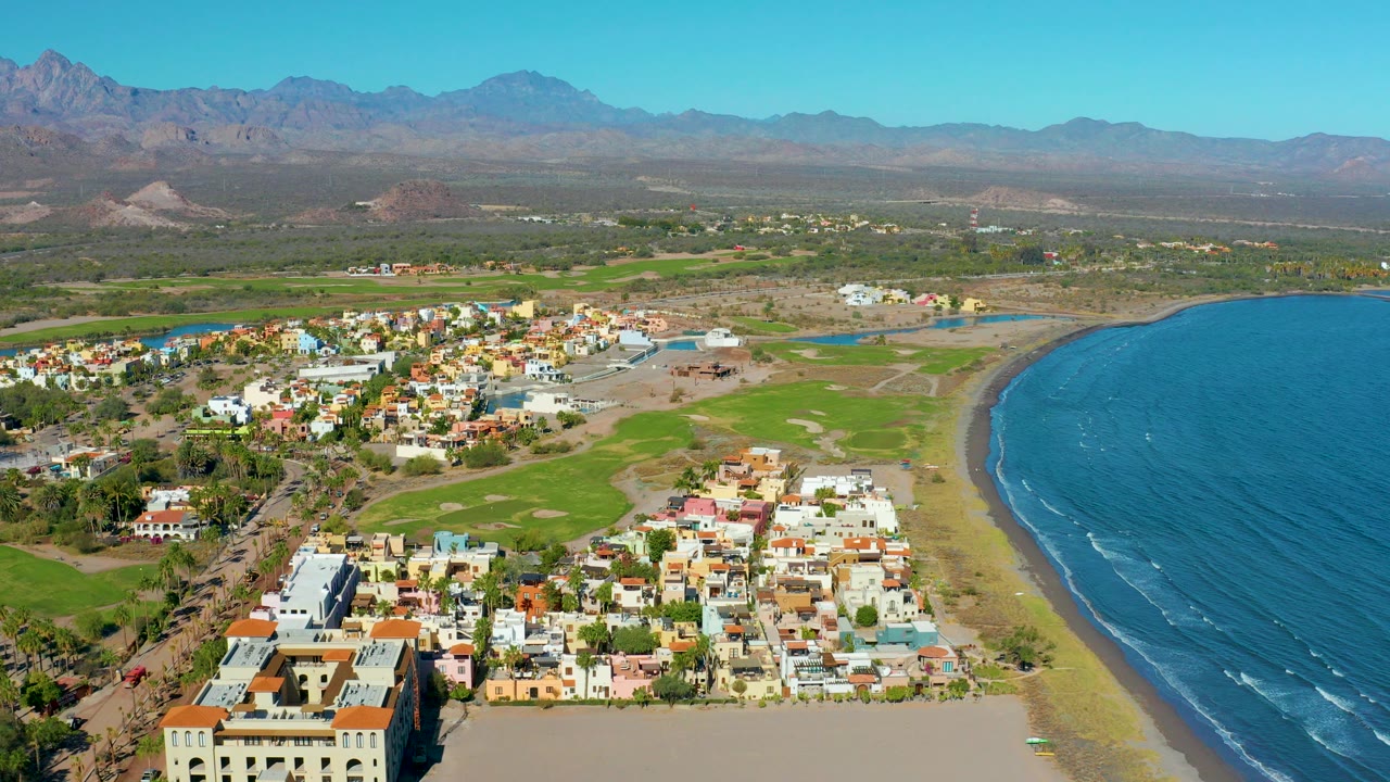 Aerial view over Loreto, Baja California Sur with colorful low-rise buildings in the foreground, a curved sandy shore and blue ocean at right, mountains beyond, and no visible people.