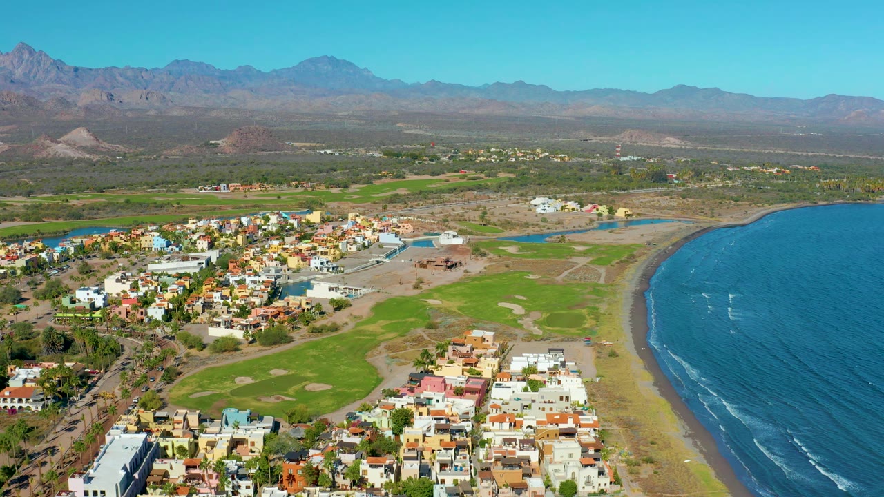 Aerial view over Loreto showing residential buildings in the foreground, green fairways near the beach, the ocean on the right, mountains across the horizon, and no visible people.