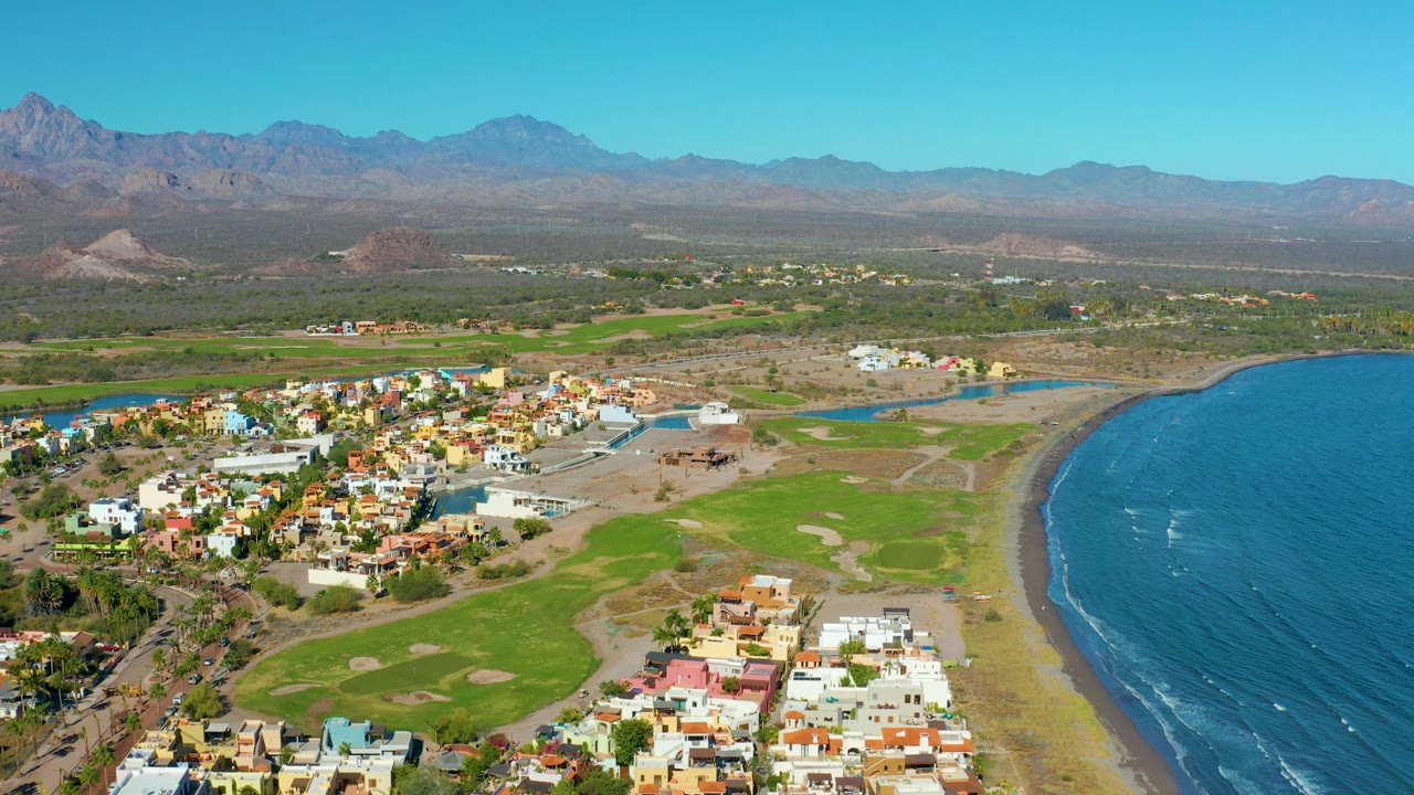 Aerial coastline scene with town, green course land, beach, ocean, and mountains.