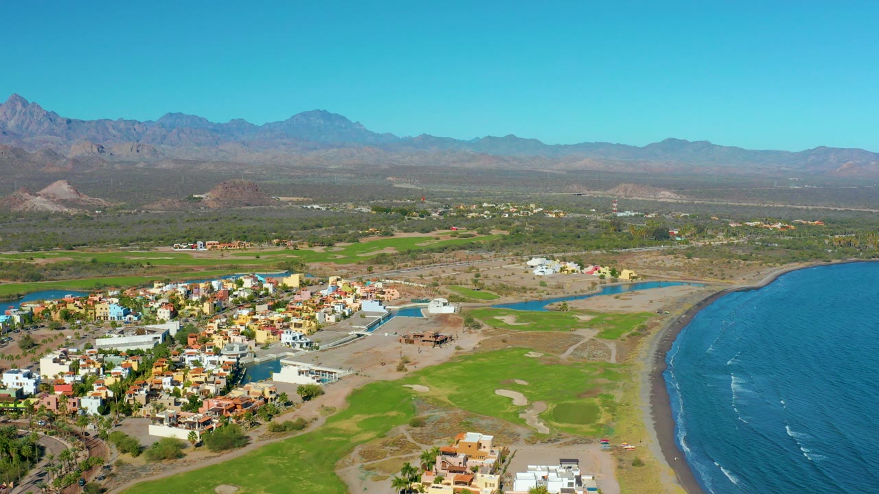 A wide aerial frame over Loreto, Baja California Sur shows colorful low-rise buildings near a narrow waterway, green ground, blue ocean at right, desert mountains on the horizon, and no visible people.