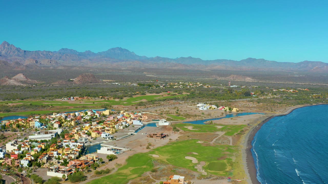 Wide aerial coastline with town, water channel, green land, and mountains.