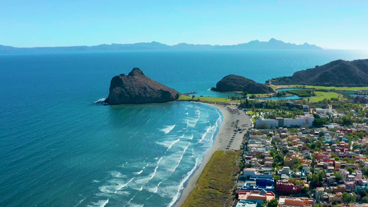 Aerial frame over Loreto, Baja California Sur showing turquoise surf on a sandy beach, low town buildings, rocky outcrops, distant mountains, and no visible people.