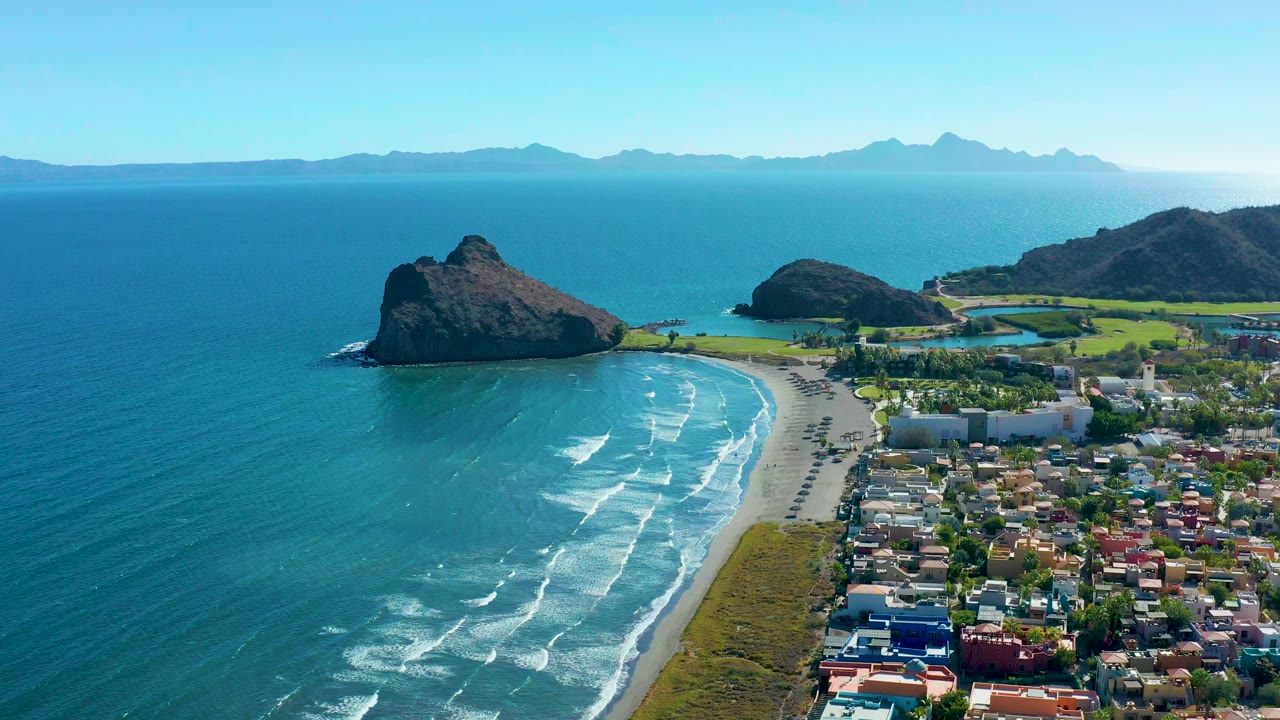 Loreto aerial frame with white wave bands advancing toward the shore, the town filling the lower right, rocky outcrops in the midground, and no visible people.