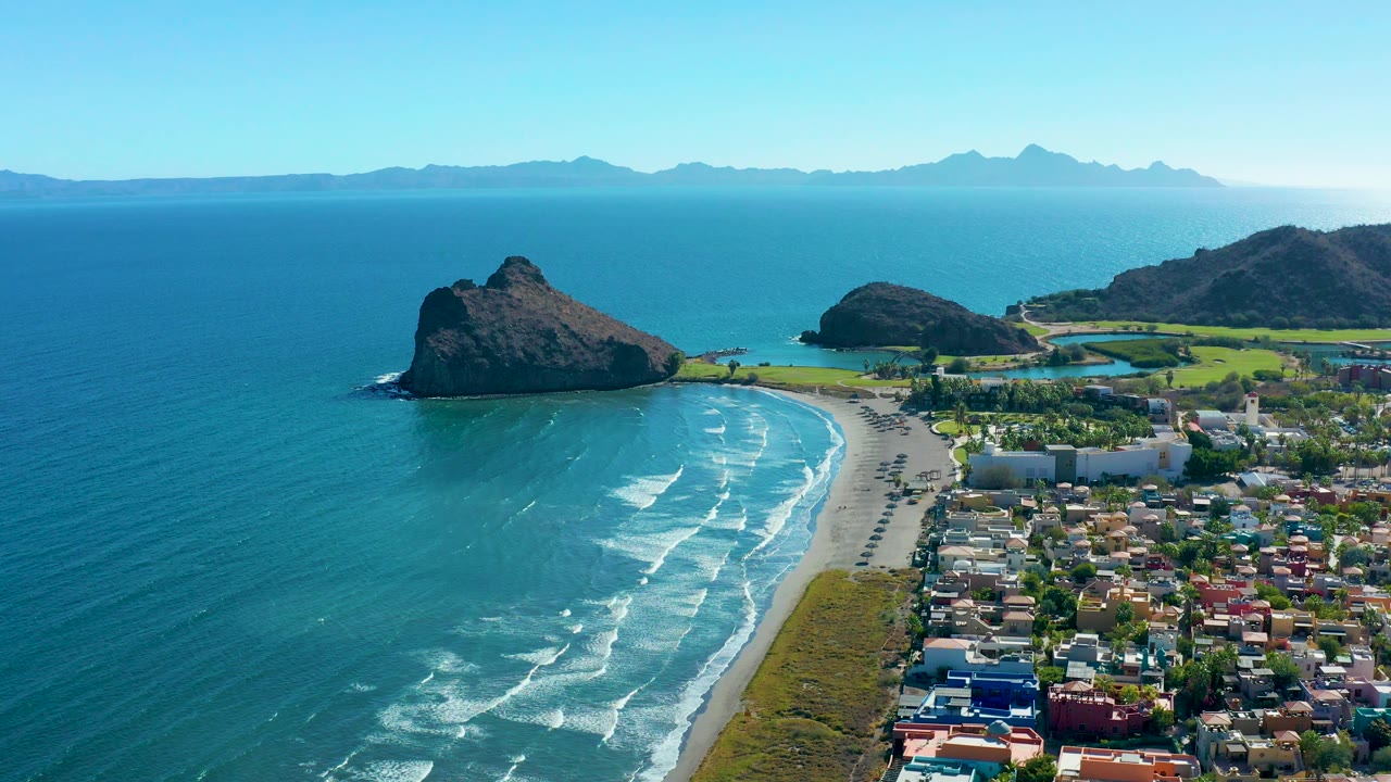Aerial coastline with blue water, white surf, sandy beach, town, and rock formations.