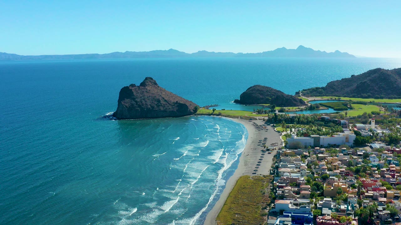 Opening aerial frame over Loreto, Baja California Sur shows turquoise ocean, a curved sandy beach, road traffic, town buildings, rocky outcrops, distant mountains, and no visible people.
