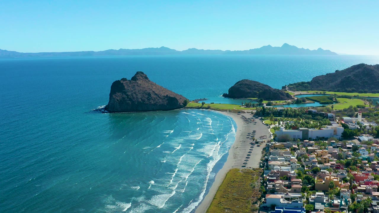 Aerial beach scene with surf, coastal road vehicles, buildings, green fields, rocks, and horizon mountains.