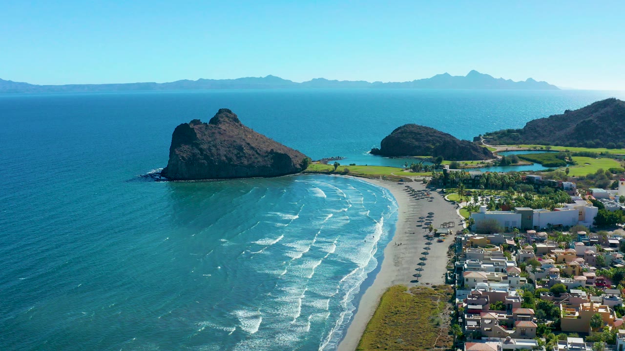 Aerial frame over Loreto, Baja California Sur showing turquoise ocean, a curved sandy beach, a coastal road, low homes, a rocky outcrop offshore, distant mountains, and no visible people.