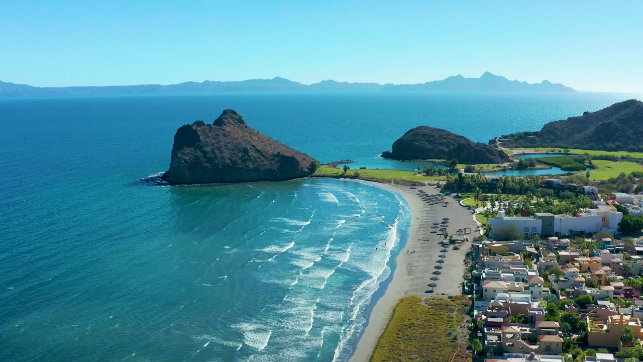 Aerial frame from Loreto, Baja California Sur showing broken wave lines across blue-green water, a pale beach, cars on the coastal road, buildings near the shore, rocky outcrops, distant mountains, and no visible people.