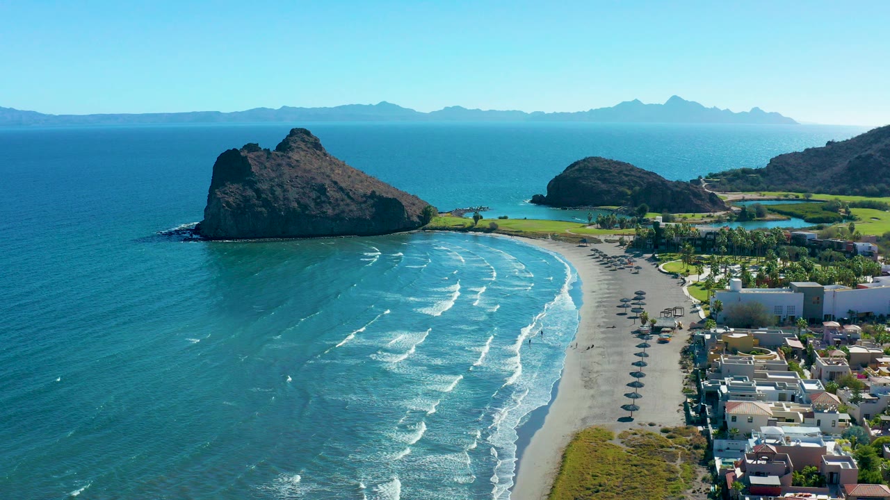 Aerial frame over Loreto, Baja California Sur, Mexico showing a curved sandy beach, blue water with white surf lines, two dark rock formations, rows of umbrellas, coastal buildings, distant mountains, and no visible people.