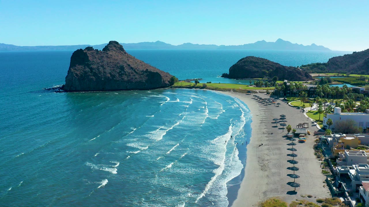 Aerial view over Loreto, Baja California Sur showing turquoise waves reaching a sandy beach, a large rocky outcrop to the left, beach umbrellas on the right, and small visible beachgoers.