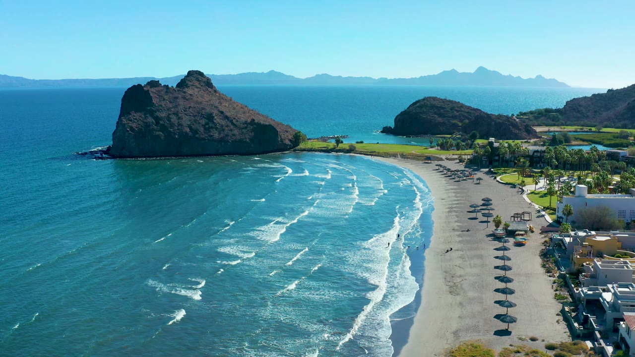 Wide coastal aerial with waves, umbrellas, buildings, and rocky landforms.