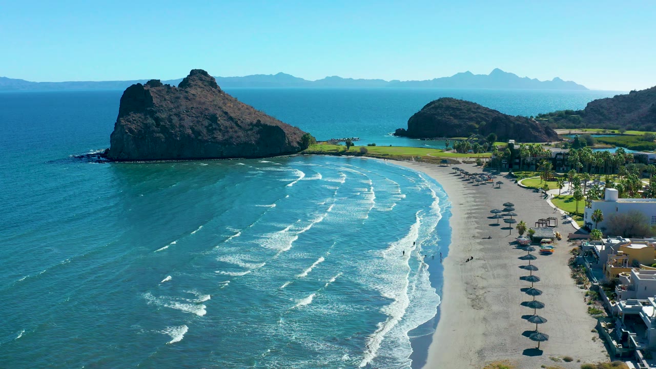 Drone-like view over Loreto, Baja California Sur with blue water filling more of the left side, wave foam moving toward the beach, a rocky outcrop near the upper left, and umbrellas arranged along the right side of the sand.