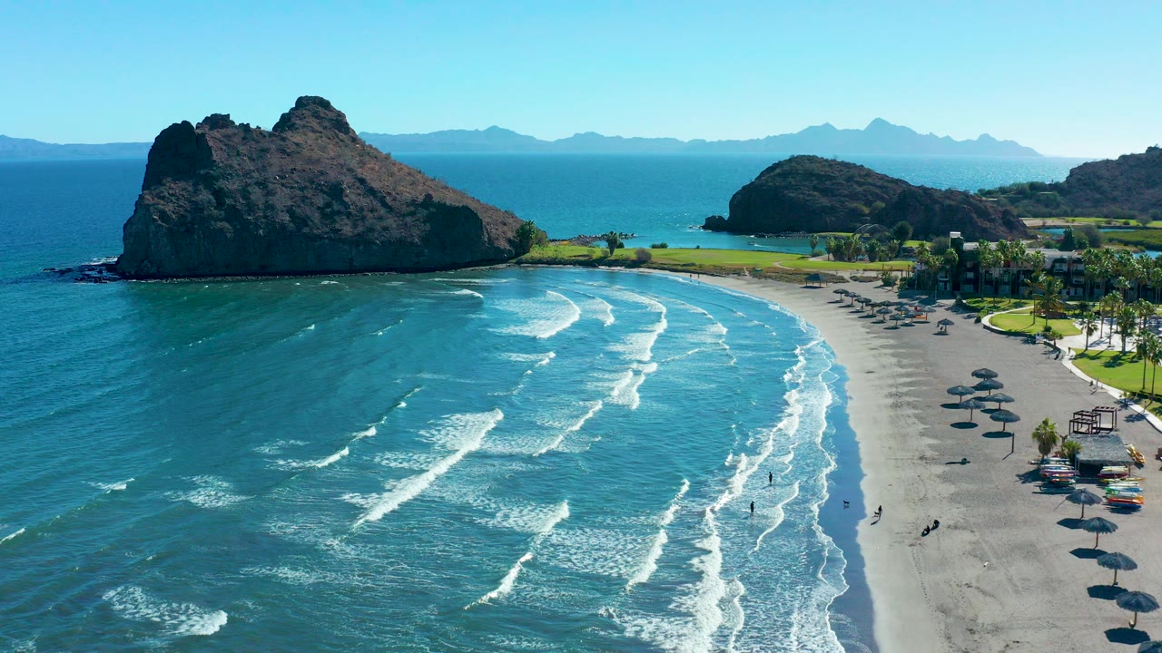 Aerial frame over Loreto, Baja California Sur, Mexico, showing white surf lines crossing blue-green water toward a sandy beach with rocky headlands in the background.