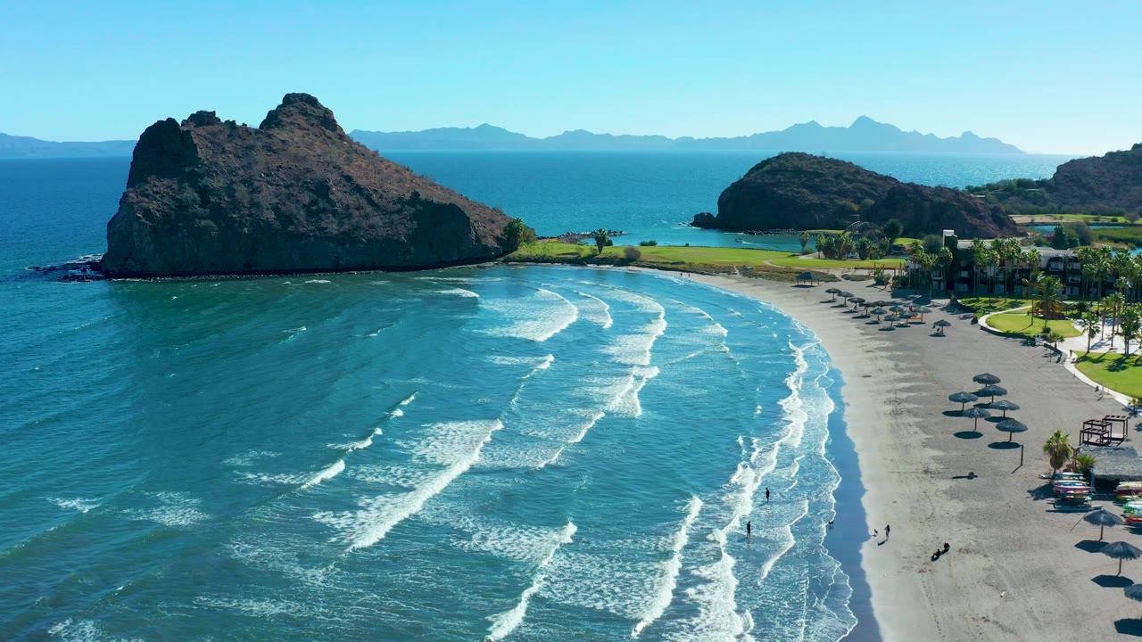 Aerial Loreto beach frame with parallel waves breaking toward the right side of the sand, umbrellas lining part of the beach, and dark rocky headlands under a clear sky.
