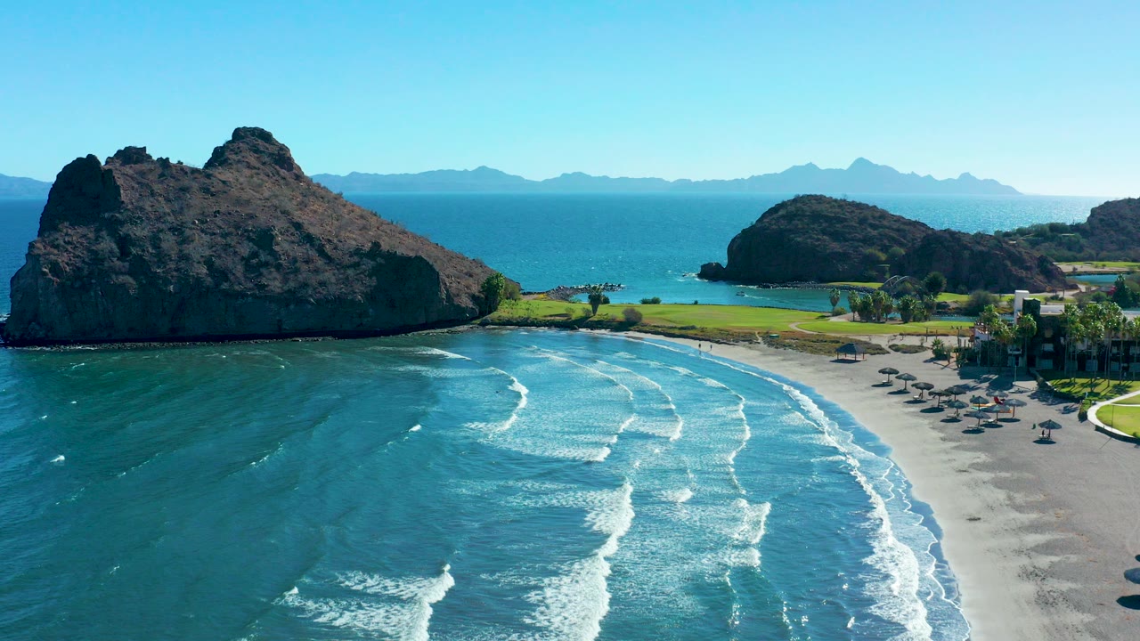 Aerial view in Loreto, Baja California Sur, with turquoise waves breaking toward a sandy beach, umbrellas on the right, dark rock formations ahead, and distant mountains under a clear sky.