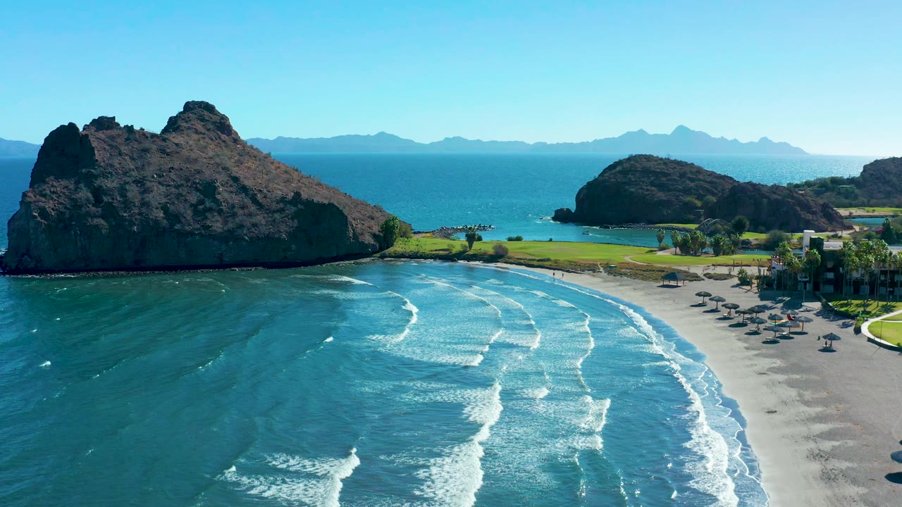 Wide aerial beach view with wave lines curving toward umbrellas and rocks.