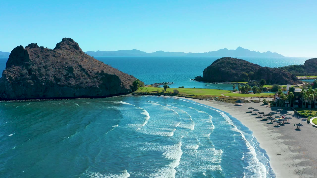 A drone-style view in Loreto places the sandy beach and active foam lines below, a large dark headland across the center, a smaller rocky point to the right, and hazy mountains beyond the water.