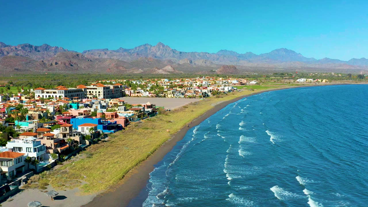 Opening aerial frame over Loreto, Baja California Sur showing colorful coastal homes, a narrow sandy beach, blue ocean with white wave lines, distant mountains, and no visible people.