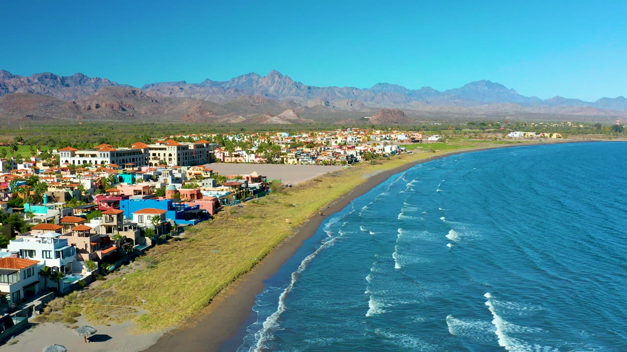 Aerial coastline with homes, green beach edge, surf, and mountain horizon.