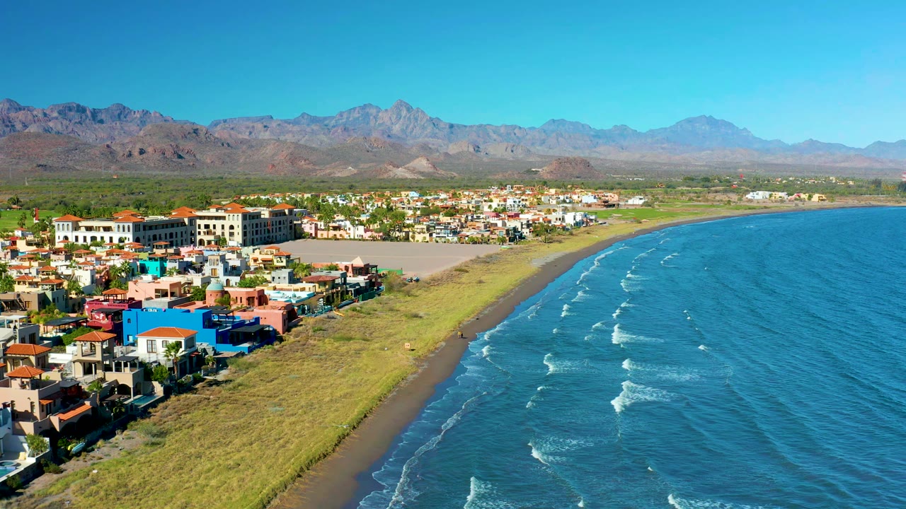 Coastal town beside a grassy beach with blue water and mountains behind.