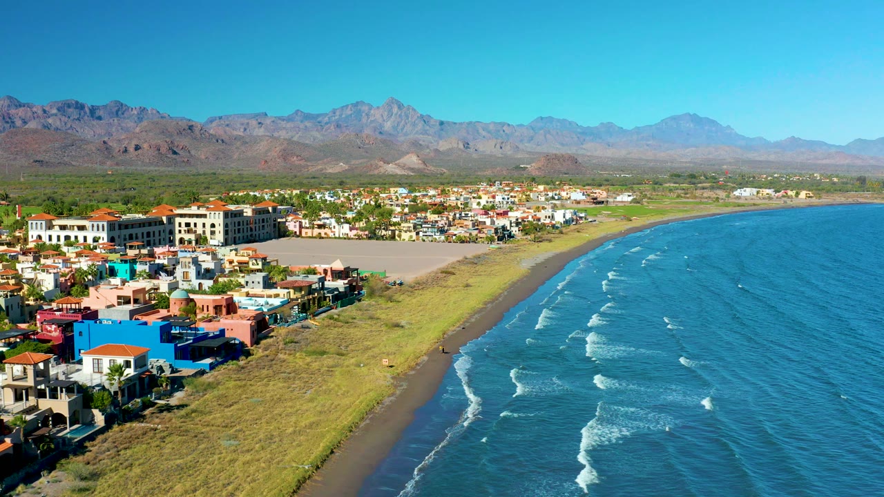Aerial shoreline view with coastal buildings, open beach, ocean, and mountains.