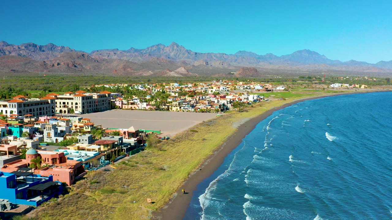 Aerial frame over Loreto, Baja California Sur showing colorful waterfront buildings at lower left, a sandy and grassy shoreline, blue ocean waves at right, distant mountains, clear sky, and no visible people.