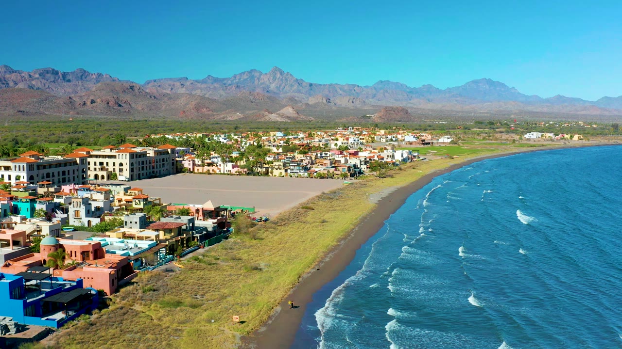 Aerial view in Loreto, Baja California Sur with low coastal buildings in the foreground, a broad sandy strip, small white wave lines on blue water, mountain ridges in the distance, and no visible people.